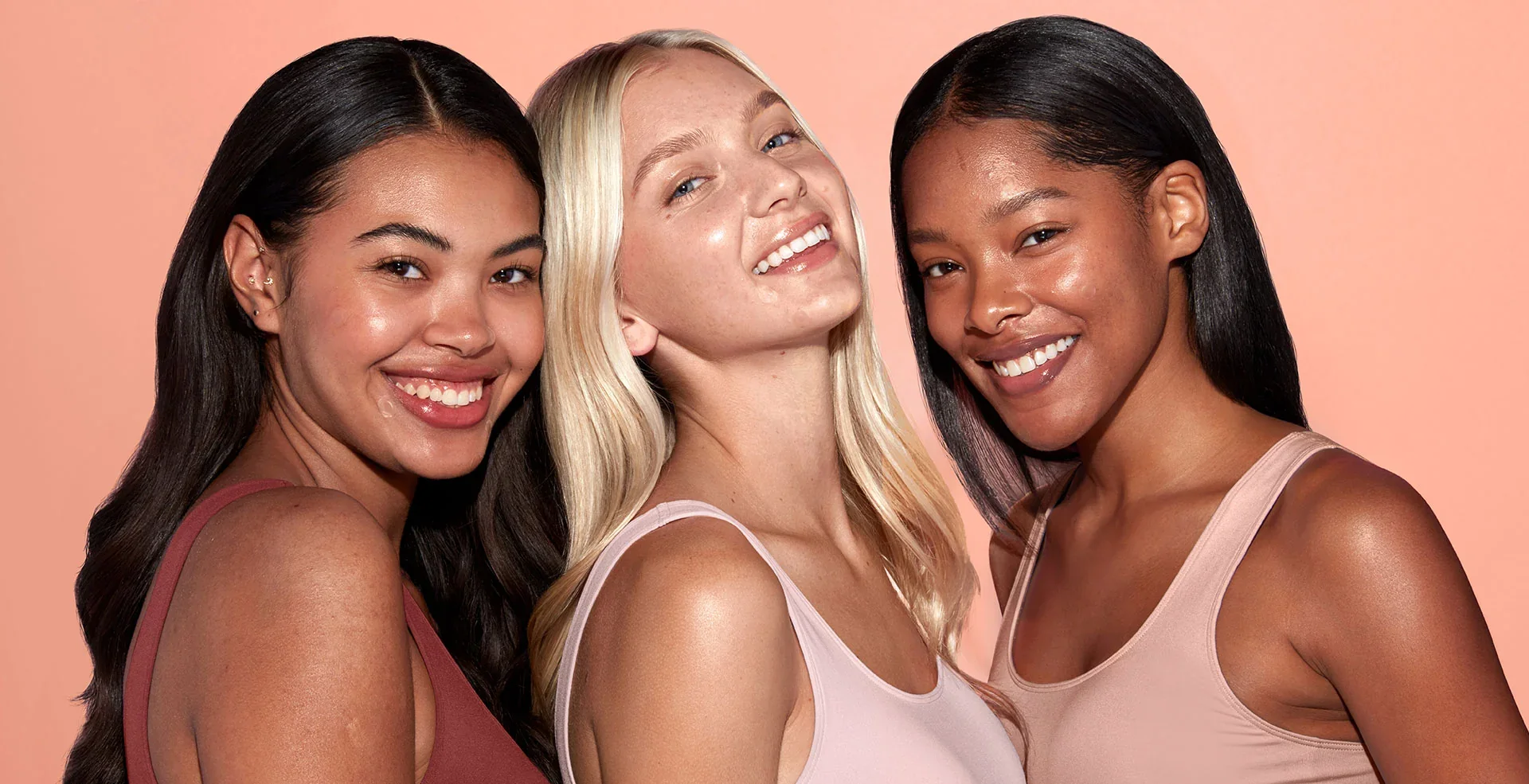 Three smiling women with glowing skin leaning together against a peach background